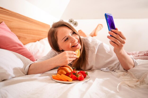 Jovem blogueiro criando conteúdo tomando selfie durante o café da manhã na cama