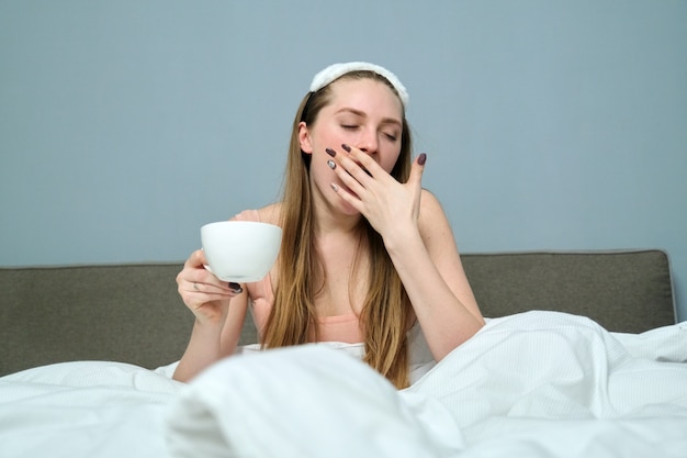 Jovem bocejando com sono na cama com uma xícara de chá