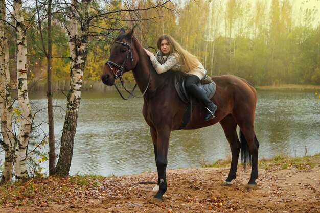 Jovem bonita a cavalo na floresta de outono perto do lago. Fêmea do cavaleiro dirige seu cavalo no parque em um clima nublado e inclemente com chuva. Conceito de passeios ao ar livre, esportes e recreação. Copie o espaço