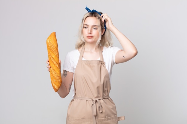 Jovem, bonita, albina, perplexa e confusa, coçando a cabeça com uma baguete de pão