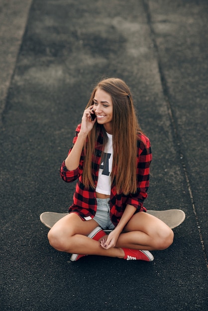 Jovem bonita bonita de camisa branca, camisa vermelha, shorts e tênis, sentado em um skate e falando no telefone celular.