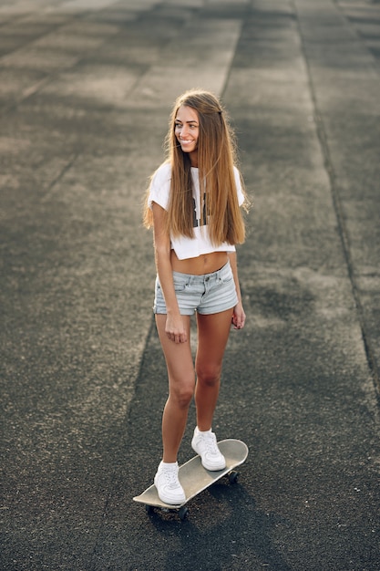 Jovem bonita bonita em uma camiseta branca, shorts curtos e tênis brancos andando de skate no estádio