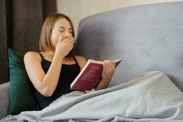 Jovem bonita cansada cobrindo a boca com a mão enquanto boceja e deitado no sofá com um livro, embrulhado em uma manta de lã, em casa. relaxe o conceito