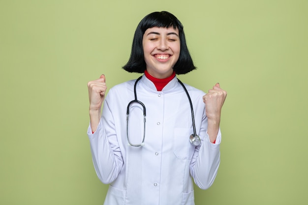 Jovem, bonita, caucasiana, satisfeita, com uniforme de médico e estetoscópio, mantendo os punhos em pé e com os olhos fechados