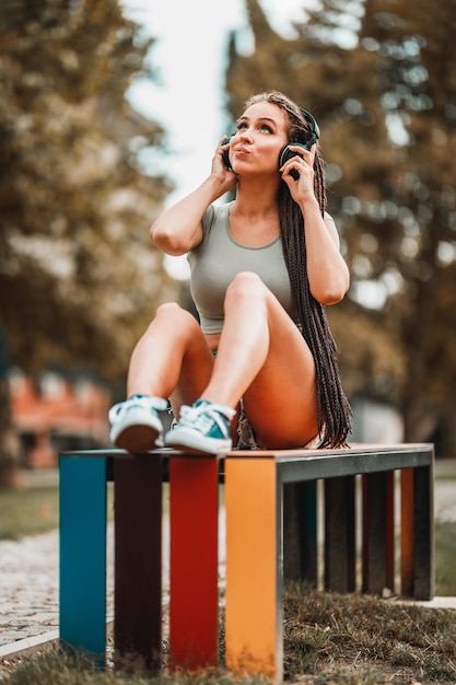 Jovem bonita com cabelo longo afro trançado ouvindo música de fones de ouvido enquanto está sentado no banco colorido do parque.