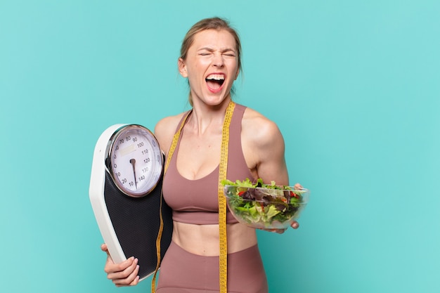 Jovem bonita com expressão de raiva de mulher esportiva segurando uma balança e uma salada