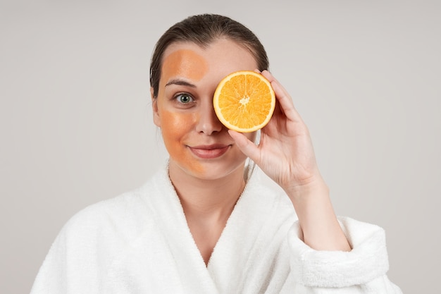 Foto jovem bonita com um manto branco aplicando uma máscara laranja no rosto