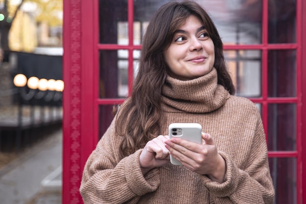 Jovem bonita com um smartphone no fundo de uma cabine telefônica vermelha.