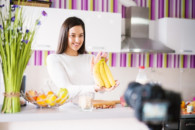 Jovem bonita e fofa está posando para uma câmera enquanto segura bananas em uma cozinha brilhante.