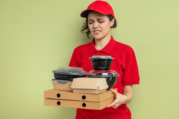 Jovem bonita entregadora descontente segurando e olhando para recipientes de comida com embalagens em caixas de pizza