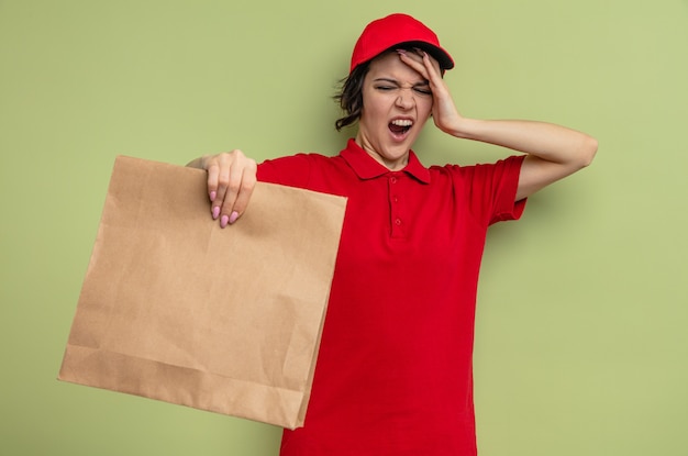 Jovem bonita entregadora irritada colocando a mão na testa e segurando uma embalagem de comida de papel