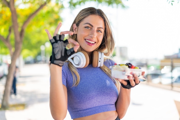 Foto jovem bonita esportiva segurando uma tigela de frutas ao ar livre mostrando sinal de ok com os dedos