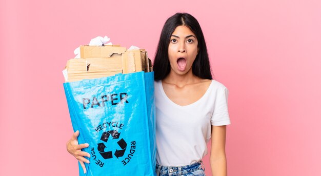 Jovem, bonita, hispânica, parecendo muito chocada ou surpresa, segurando um saco de papel para reciclar