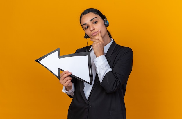 Jovem bonita impressionada usando um blazer preto com fone de ouvido segurando a marca de direção e colocando a mão no queixo