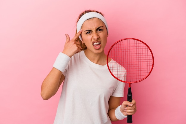 Jovem bonita loira caucasiana segurando uma raquete de badminton isolada na parede rosa, mostrando um gesto de decepção com o dedo indicador