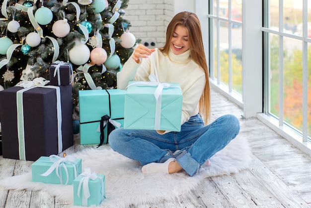 jovem bonita perto da árvore de natal abre presentes de natal em uma sala bem iluminada e aconchegante