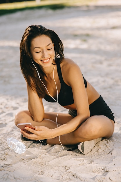 Jovem bonita sorrindo moda bronzeada estilo desportivo mulheres vestidas de biquíni preto, sentado em uma praia