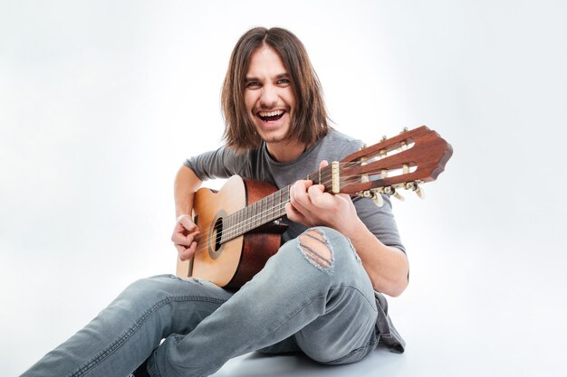 Foto jovem bonito alegre com cabelo comprido sentado e tocando guitarra sobre um fundo branco