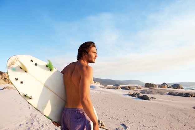 Jovem bonito andando na praia com prancha de surf