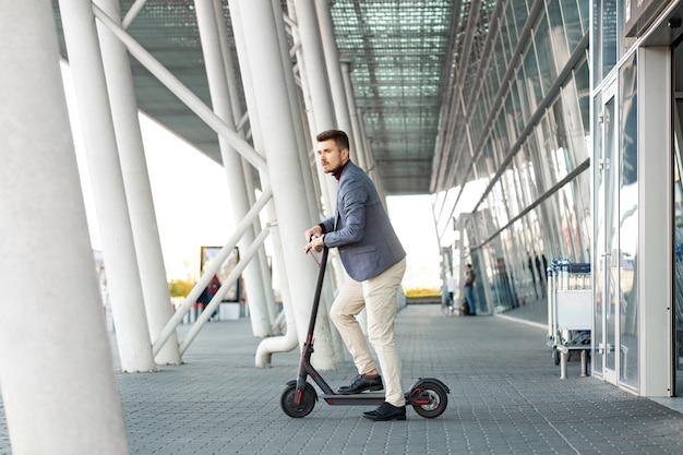 Jovem bonito andar e-scooter sai do aeroporto. alugue um transporte em uma viagem. transporte urbano na moda na moderna scooter elétrica. conceito de mobilidade amigável de eco.