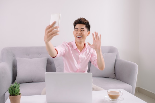 Jovem bonito asiático com laptop, tirando uma selfie com o telefone, sentado em um sofá em casa