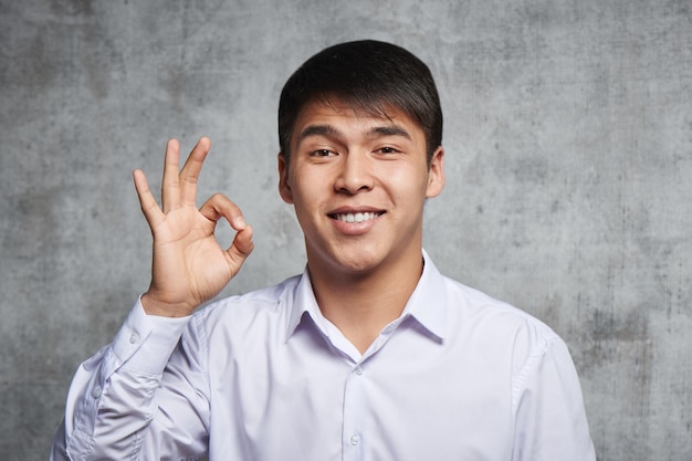 Jovem bonito asiático mostra aos dedos um gesto de que está tudo bem. Cazaque feliz alegre em roupas de estilo empresarial em fundo cinza um