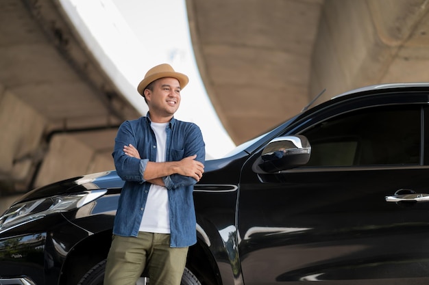 Jovem bonito asiático recebendo o carro novo. Ele está de pé com o braço cruzado na frente do carro. Ele está muito feliz. Compre ou alugue um conceito de carro.