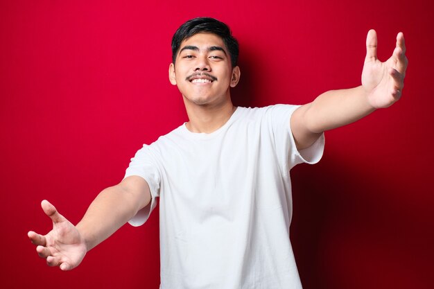 Jovem bonito asiático vestindo camiseta branca casual, olhando para a câmera, sorrindo com os braços abertos para um abraço. expressão alegre abraçando a felicidade sobre fundo vermelho