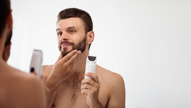 Jovem bonito barbear a barba no banheiro. Retrato de um homem barbudo nu elegante, examinando seu espelho em casa de rosto.