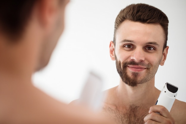 Jovem bonito barbear a barba no banheiro. Retrato de um homem barbudo nu elegante, examinando seu espelho em casa de rosto.