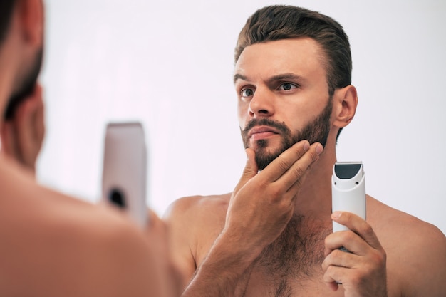 Jovem bonito barbear a barba no banheiro. Retrato de um homem barbudo nu elegante, examinando seu espelho em casa de rosto.