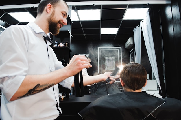 Jovem bonito barbeiro fazer corte de cabelo de menino bonitinho na barbearia.