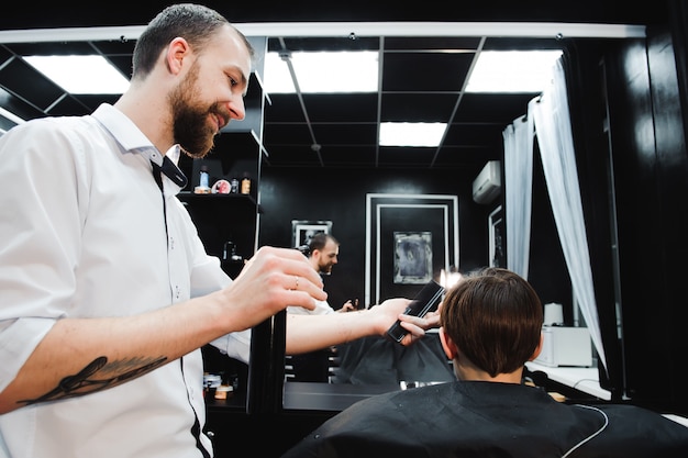 Jovem bonito barbeiro fazer corte de cabelo de menino bonitinho na barbearia.