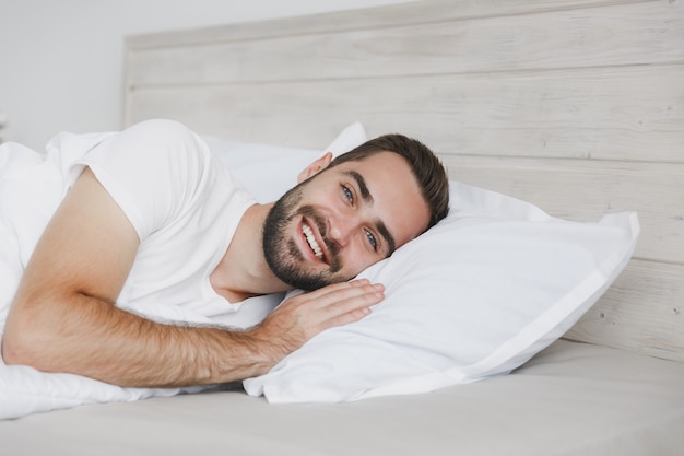 Jovem bonito barbudo calmo deitado na cama com lençol branco e cobertor no quarto em casa
