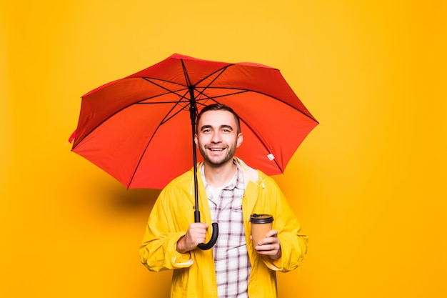 Jovem bonito barbudo com capa de chuva amarela e guarda-chuva vermelho bebendo café isolado sobre fundo laranja