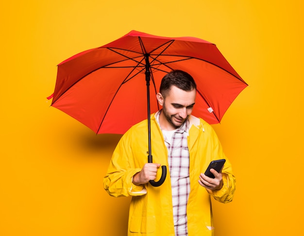 Jovem bonito barbudo com capa de chuva amarela e guarda-chuva vermelho usando telefone celular isolado sobre fundo laranja