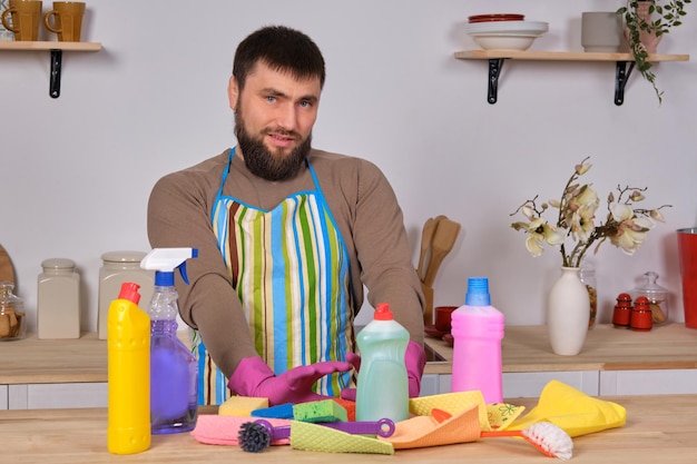 Jovem bonito barbudo na cozinha, mostra toda a sua equipe de limpeza - detergentes, escovas, sprays. Ele acha que está pronto para uma limpeza real