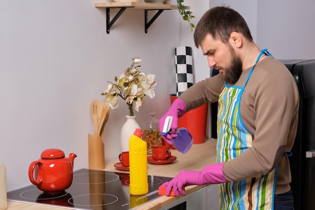 Jovem bonito barbudo na cozinha, usando avental e luvas cor-de-rosa, limpa a superfície da cozinha com detergentes