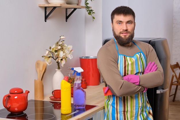 Jovem bonito barbudo na cozinha, usando avental e luvas cor-de-rosa, limpa a superfície da cozinha com detergentes