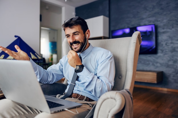 Jovem bonito barbudo pai vestido elegante sentado na cadeira em casa e usando o laptop para chamada em conferência.