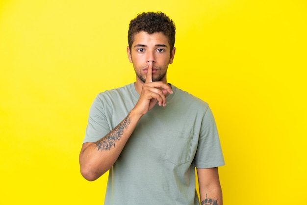 Jovem bonito brasileiro isolado em fundo amarelo mostrando sinal de silêncio gesto de colocar o dedo na boca