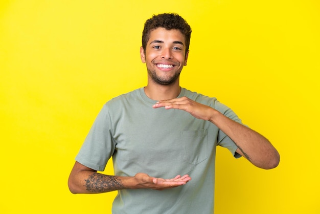 Jovem bonito brasileiro isolado em um fundo amarelo segurando copyspace imaginário na palma da mão para inserir um anúncio