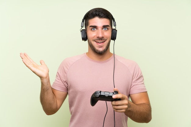 Jovem bonito brincando com um controlador de videogame sobre parede verde isolada com expressão facial chocada