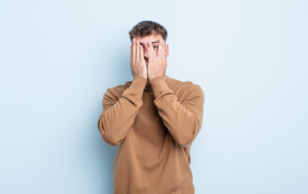 Jovem bonito cobrindo o rosto com as mãos, espiando por entre os dedos com expressão de surpresa e olhando para o lado