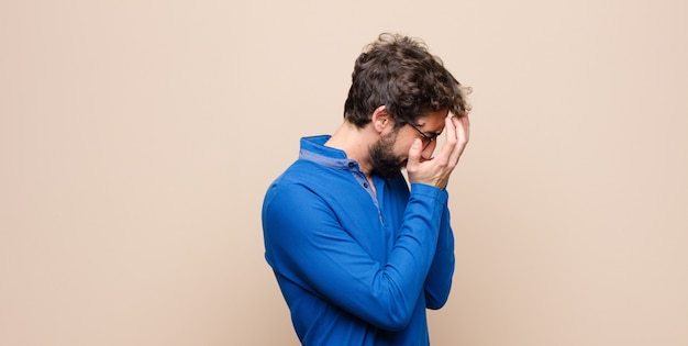 Jovem bonito cobrindo os olhos com as mãos com um olhar triste e frustrado de desespero, chorando, vista lateral em parede plana