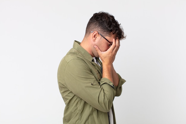 Jovem bonito cobrindo os olhos com as mãos com uma expressão triste e frustrada de desespero, chorando, vista lateral