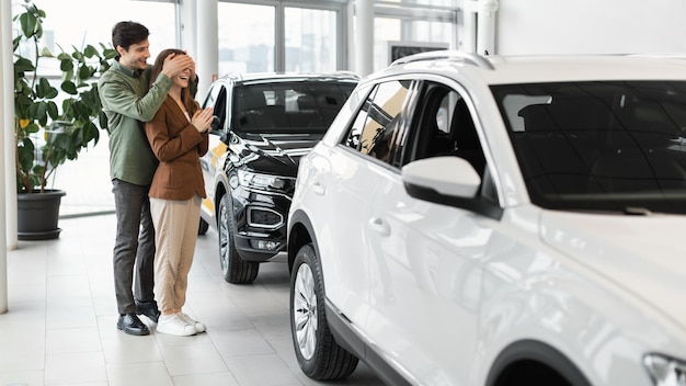 Jovem bonito cobrindo os olhos da esposa fazendo presente surpresa comprando carro novo no automóvel