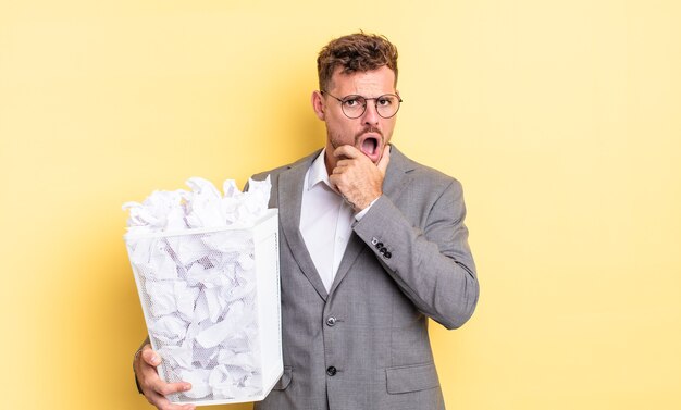 Jovem bonito com a boca e os olhos bem abertos e a mão no queixo conceito de lixo de bolas de papel