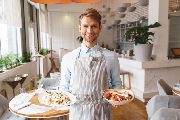 Jovem bonito com avental olhando e sorrindo enquanto segura deliciosas saladas frescas