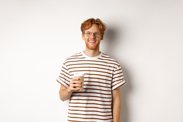 Jovem bonito com barba e cabelo ruivo bagunçado, usando óculos com camiseta listrada, tomando café para viagem e sorrindo, fundo branco.